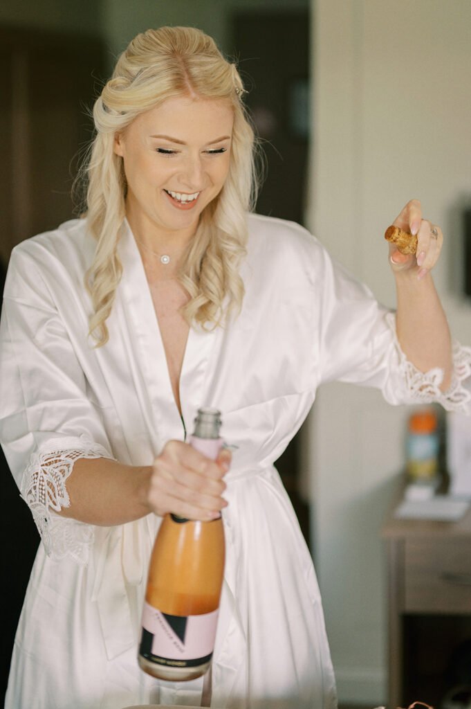 A bride to be with long blonde hair, wearing a white robe, joyfully opens a bottle of champagne. Captured by a Coniston Hotel wedding photographer, she holds the cork in one hand and the bottle in the other, standing in an indoor setting with a beautifully blurred background.