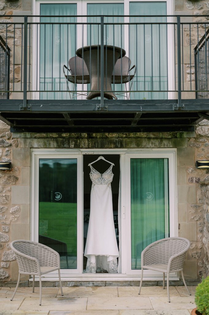 A white wedding dress hangs in a doorway beneath a stone balcony with a metal railing, perfect for that Coniston Hotel wedding vibe. The balcony features a round table and chairs, while below, two wicker chairs flank the entrance. Through the glass doors, green grass stretches invitingly.