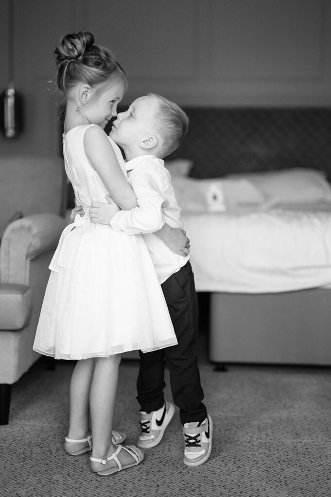 In a black and white photo capturing pure joy, a girl in a white dress with her hair in a bun and a boy in shirt, trousers, and sneakers share a playful hug. Its as if theyve momentarily turned the room—with its bed and couch—into their kingdom. A snapshot worthy of any Coniston Hotel wedding photographers portfolio.