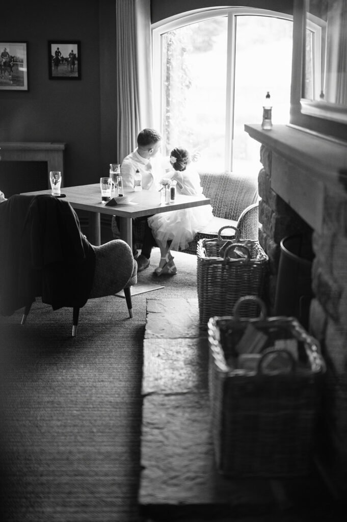 A black-and-white photo of a young boy and girl sitting by a window at a table in a cozy room. They appear to be siblings or friends, with the boy whispering to the girl, reminiscent of candid moments captured by a Coniston Hotel wedding photographer. The room has a stone fireplace and wicker baskets on the floor.