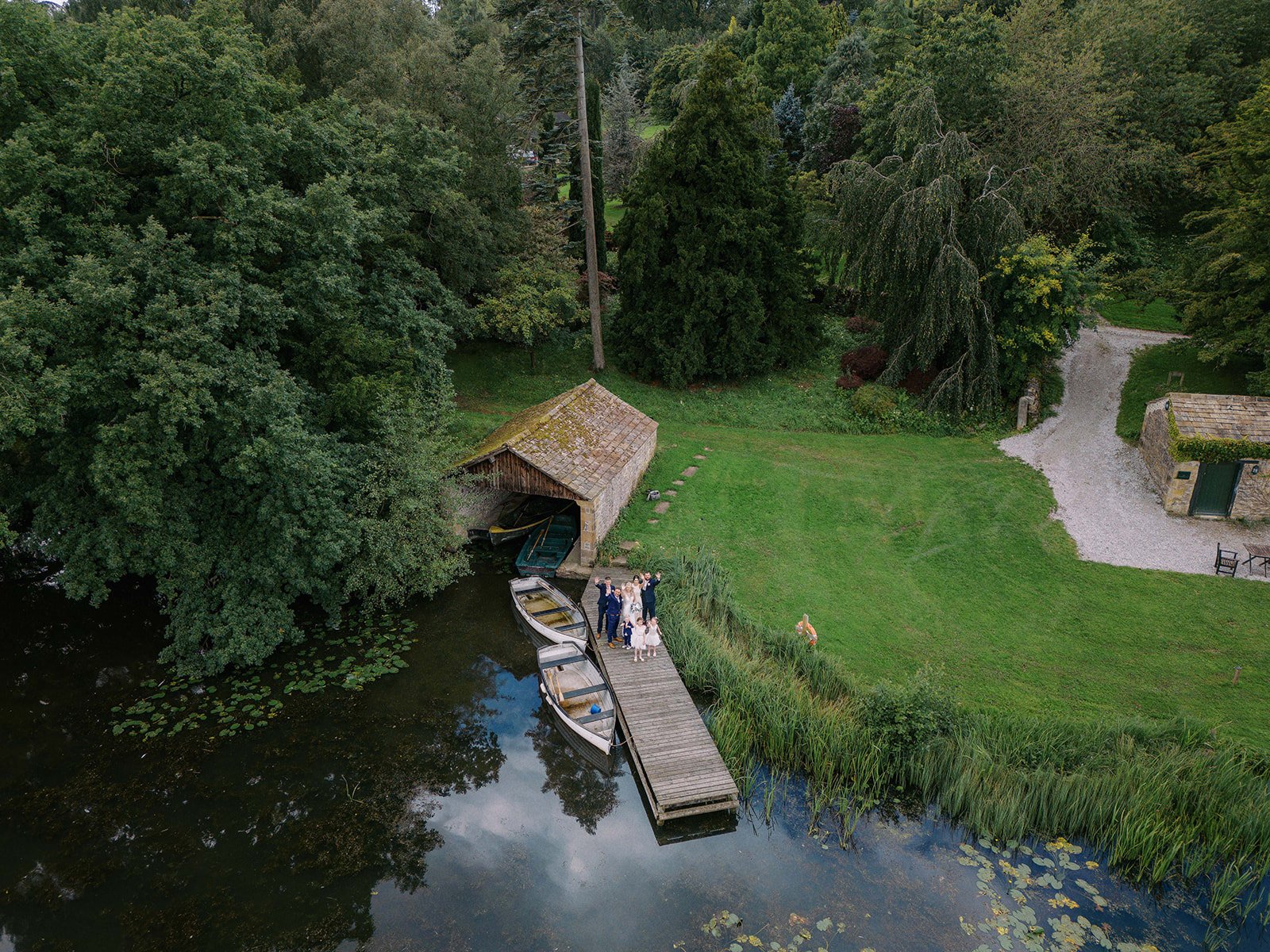 Aerial view of a serene lakeside scene near Coniston Hotel, featuring a small wooden dock and two boats. Trees embrace a rustic boathouse, where a couple with their dog stands. Lush greenery and lily pads embellish the waters edge, as a winding path leads into the trees—a perfect spot for wedding photographers.