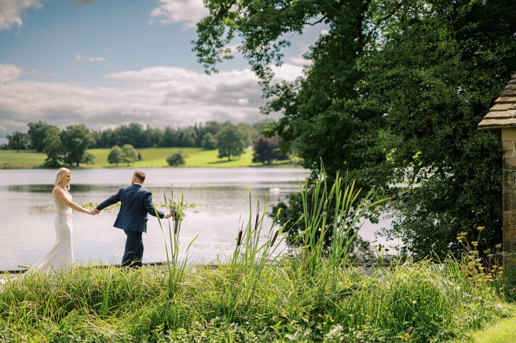 A couple in wedding attire walks hand-in-hand beside a tranquil lake at Coniston Hotel. The groom, in a dark suit, leads the bride, in a white gown. Lush greenery and tall grass surround them, with trees dotting the far shore under a partly cloudy sky—a perfect scene for a wedding photographer.