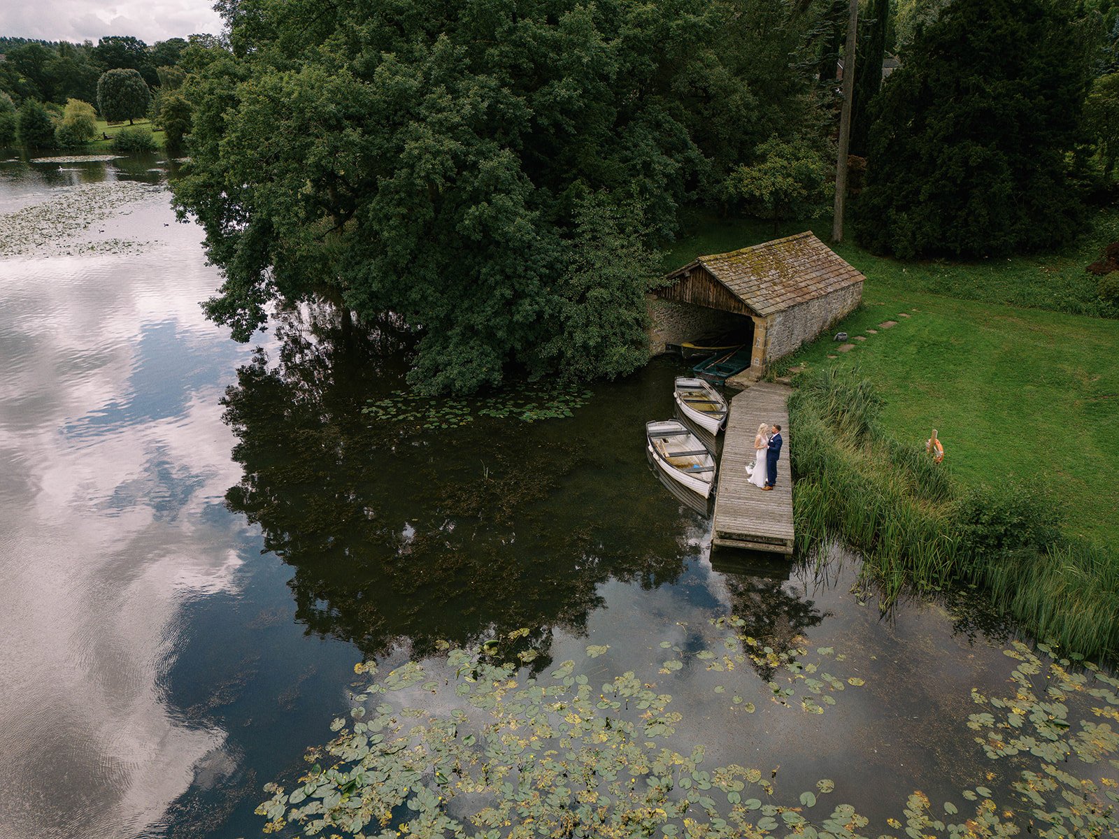 An aerial view captures a lush lakeside scene at Coniston Hotel, showcasing a wooden dock with three small boats tied to it beside a rustic boathouse. A couple stands on the dock, surrounded by grass, trees, and water lilies, making it an ideal spot for a wedding photographer.