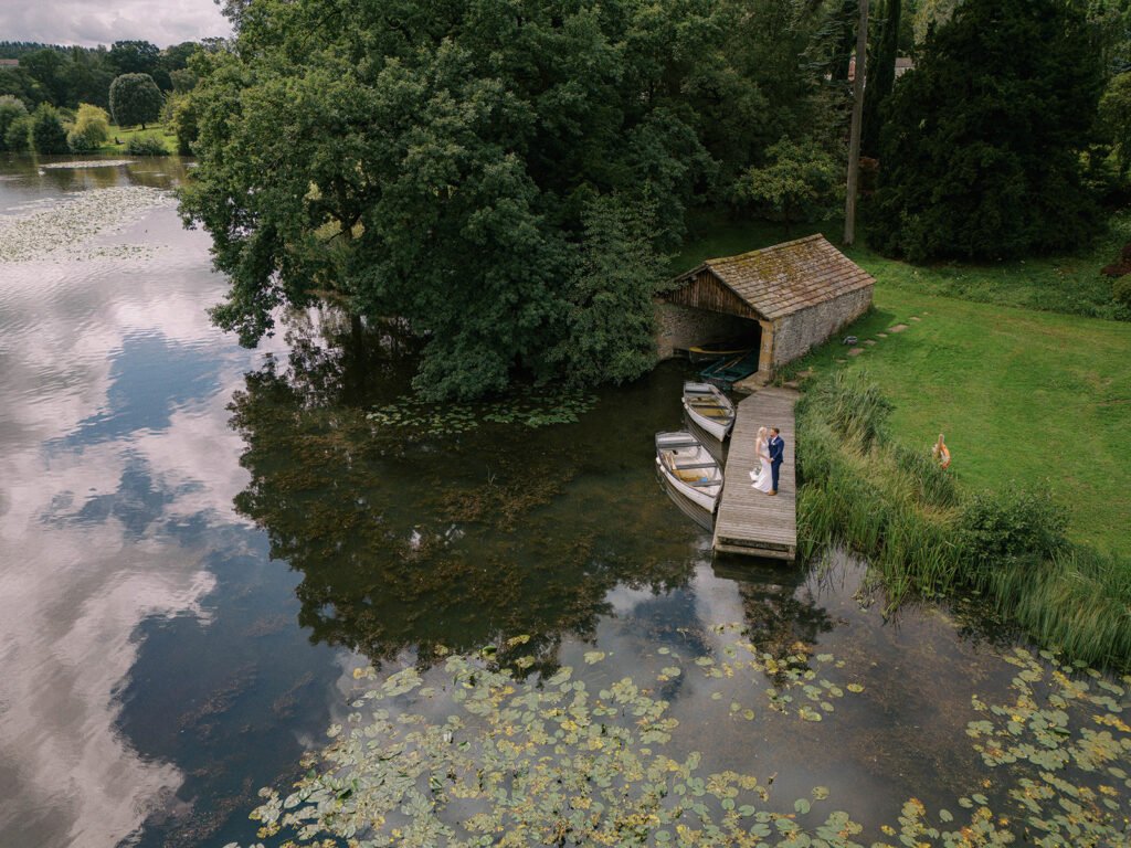 A couple stands on a wooden dock next to a small boathouse by a serene lake, captured by a Coniston hotel wedding photographer. Surrounded by lush trees and lily pads, the cloudy sky reflects in the water as a deer grazes on the grassy shore nearby.