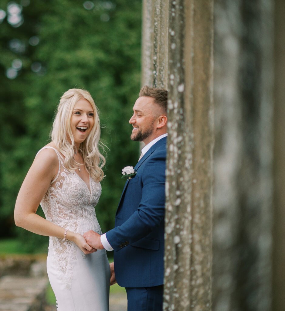 A couple in wedding attire stands outdoors, holding hands and smiling at each other. The bride wears a white lace gown, and the groom is in a blue suit with a white boutonniere, captured beautifully by a Coniston Hotel wedding photographer amid elegant stone pillars and lush greenery.