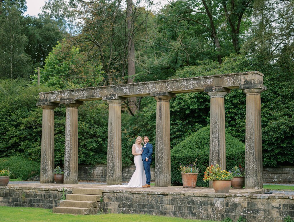 A bride in a white gown and a groom in a blue suit stand under an ancient stone colonnade at the Coniston Hotel. Amidst lush greenery and potted plants, their love story unfolds on this cloudy day, perfectly captured by their wedding photographer.