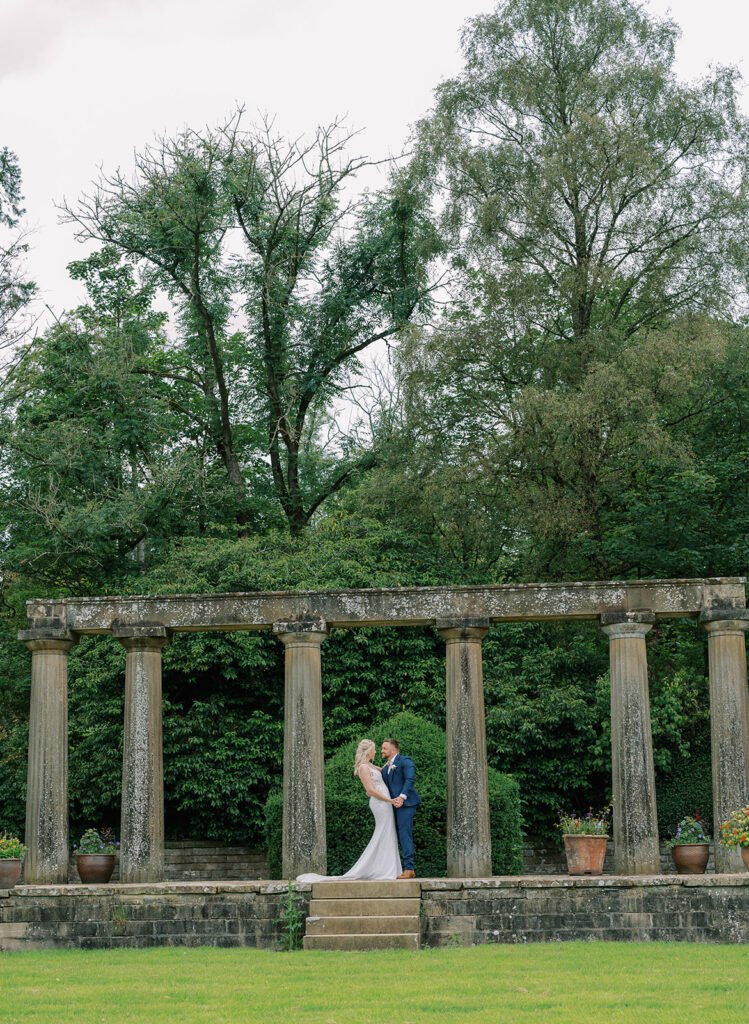 A couple in wedding attire shares an embrace on stone steps, framed by ancient columns, their moment expertly captured by a Coniston Hotel wedding photographer. Lush green trees and a well-maintained lawn provide the perfect backdrop for this enchanting scene.