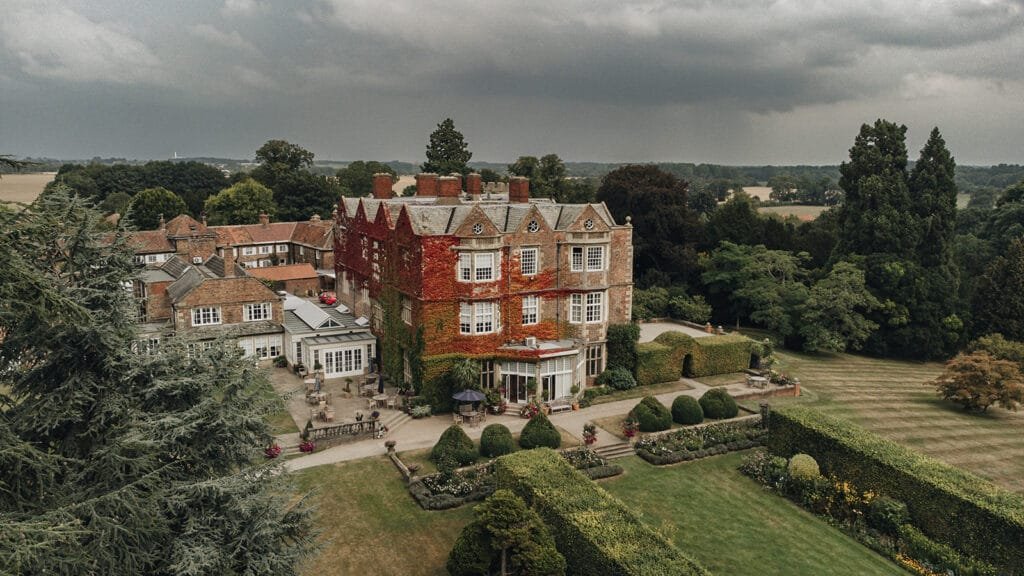 An aerial view captures the majesty of a large, historic stone mansion, its ivy-covered walls whispering tales of old. Surrounded by well-manicured gardens and lush trees under a cloudy sky, this scene is a dream canvas for any Goldsborough Hall wedding photographer seeking countryside charm.