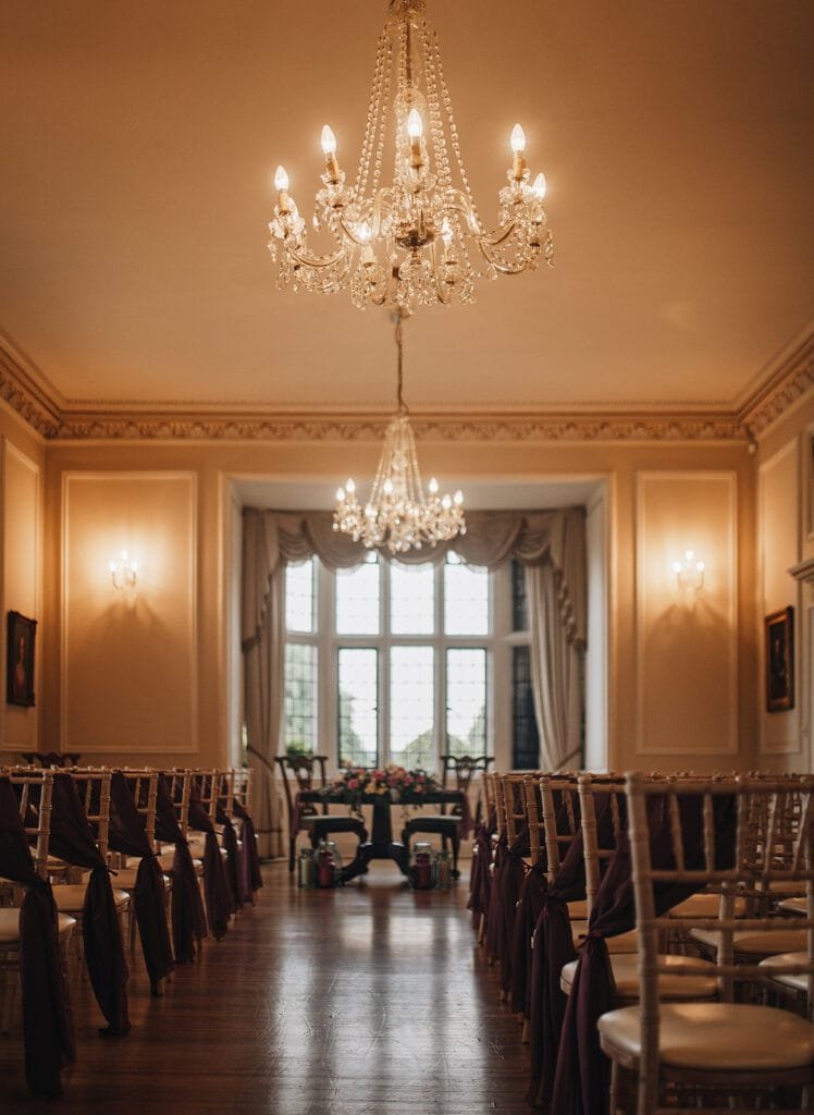 An elegant, warmly lit room at Goldsborough Hall is set up for a wedding ceremony. Rows of chairs with dark fabric sashes face a table adorned with flowers beneath ornate chandeliers. Tall windows in the background allow natural light to filter in, perfect for capturing on camera by the wedding photographer.