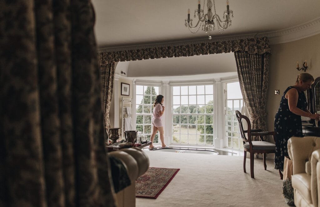 A woman in a pink dress stands near large windows at Goldsborough Hall, gazing outside. Nearby, another person leans over a chair. The rooms elegant curtains and chandelier create a warm glow, complemented by the views captured by the wedding photographer through the windows.