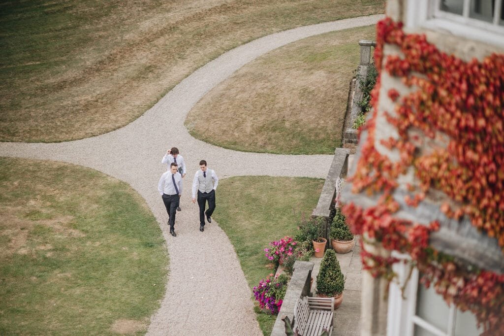 Three men in formal attire stroll along a gravel path cutting through a grassy lawn. To the right, a building draped in red ivy and adorned with colorful potted flowers adds charm to the scene—captured beautifully by a Goldsborough Hall wedding photographer. Dry patches dot the expansive green below.