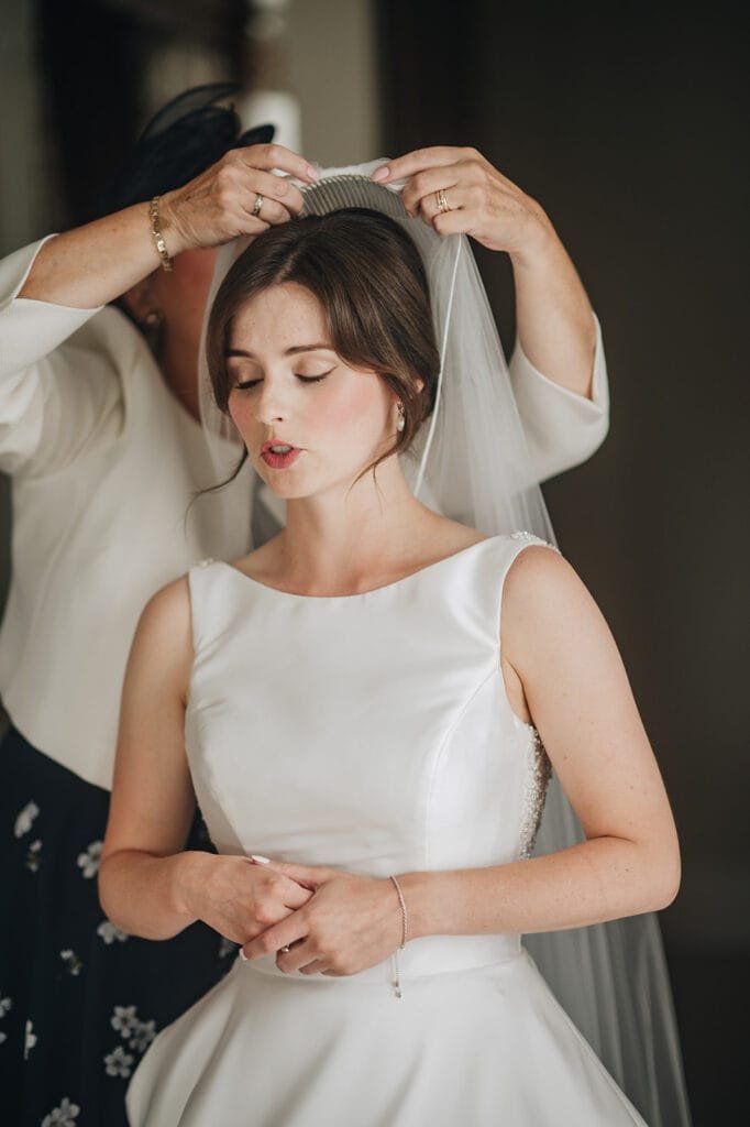 At Goldsborough Hall, a bride in a white wedding dress stands with her eyes closed as another person adjusts her veil. Her hair is styled in a low updo. The rooms soft lighting, captured by the wedding photographer, emphasizes a calm and intimate moment.