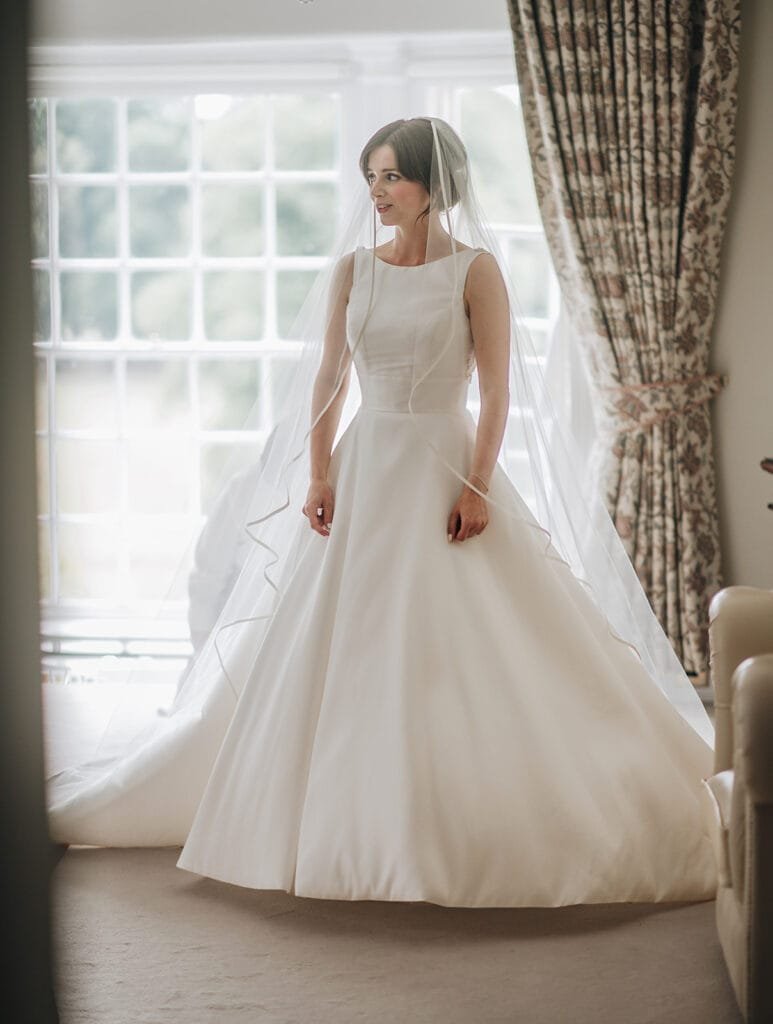 A bride in a white wedding dress stands near a large window at Goldsborough Hall. Her sleeveless gown features a full skirt and she wears a long veil. The room, perfect for capturing by a wedding photographer, boasts patterned curtains and soft lighting.