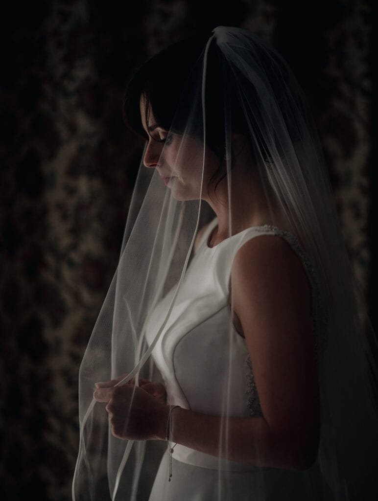 A bride in a white dress stands in profile, holding her veil. The dim lighting creates a soft, intimate atmosphere as she gazes downward. Captured by a Goldsborough Hall wedding photographer, the moment exudes timeless elegance and grace.