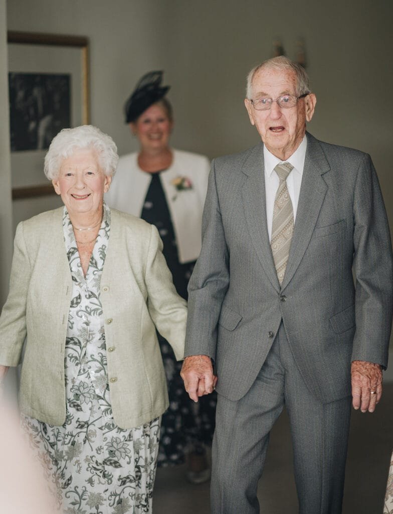 An elderly couple, dressed in elegant formal attire, hold hands and smile warmly. Behind them, a woman in a white jacket and black hat beams with joy. The indoor setting, captured by a Goldsborough Hall wedding photographer, features soft lighting and a framed picture on the wall.