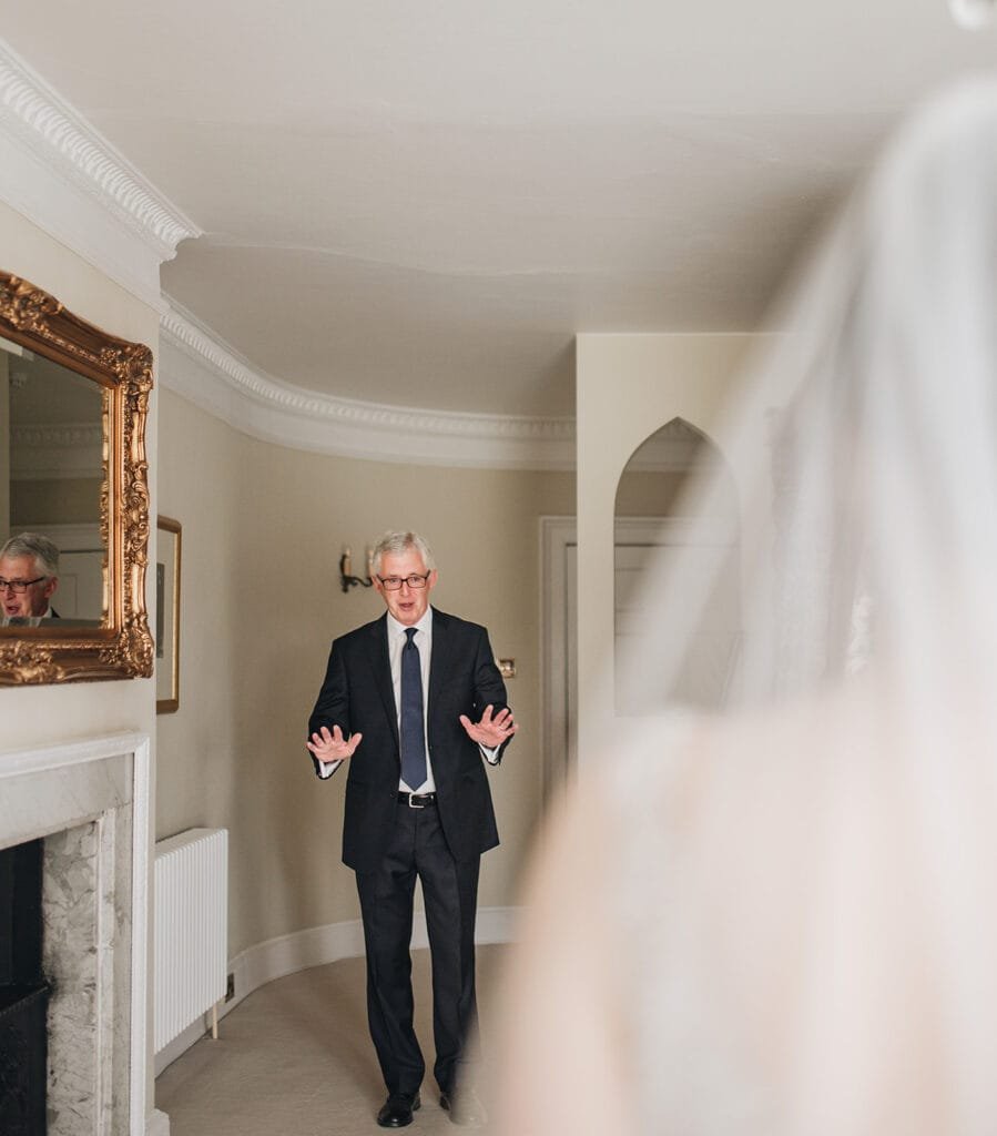 A man in a suit looks surprised and pleased, his hands raised slightly as he stands near a mirror with ornate molding. The blurred foreground hints at someone in a bridal gown approaching, captured beautifully by the Goldsborough Hall wedding photographer.