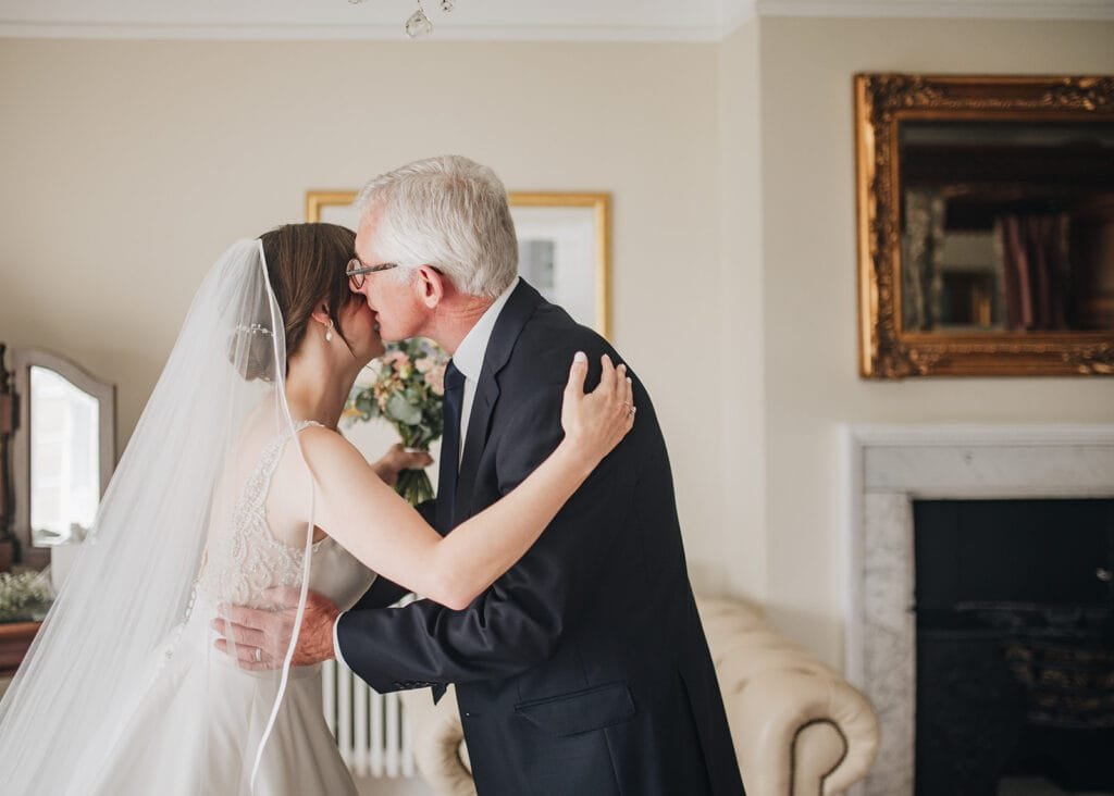 A bride in a white dress and veil embraces an older man, likely her father, in a warmly lit room at Goldsborough Hall. The scene is beautifully framed with artwork and a fireplace, captured perfectly by the wedding photographer.