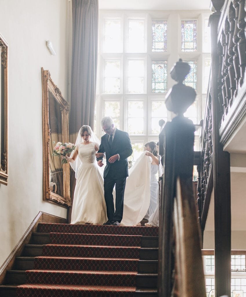 A bride in a white gown gracefully descends the staircase at Goldsborough Hall, an older man by her side. She clutches a bouquet as attendants adjust her veil. With large windows and framed paintings lining the carpeted stairs, this moment is beautifully captured by the wedding photographer.