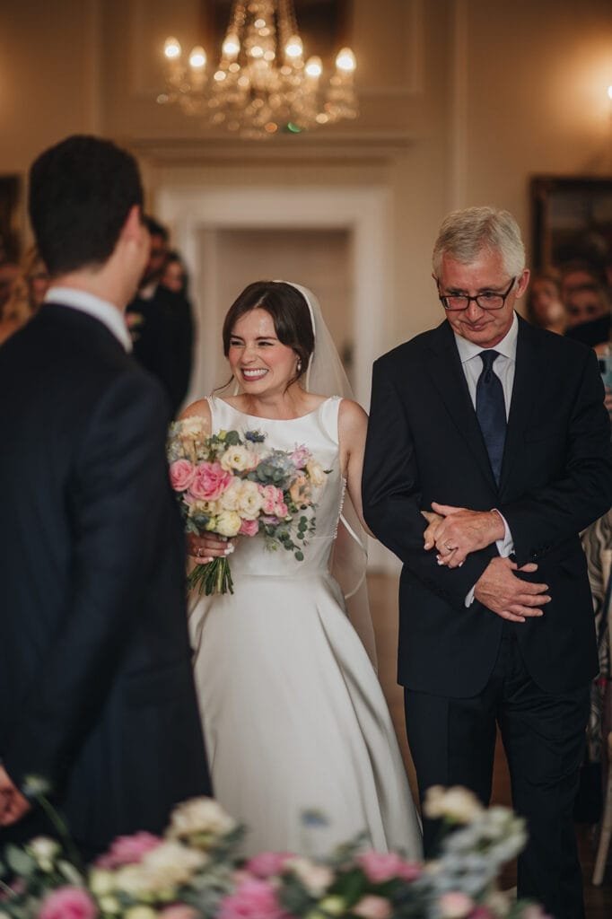 A bride in a white dress with a veil holds a colorful bouquet as she walks down the aisle at Goldsborough Hall with an older man. They smile at the groom, who stands with his back to the camera, while a chandelier elegantly illuminates the moment captured by their wedding photographer.