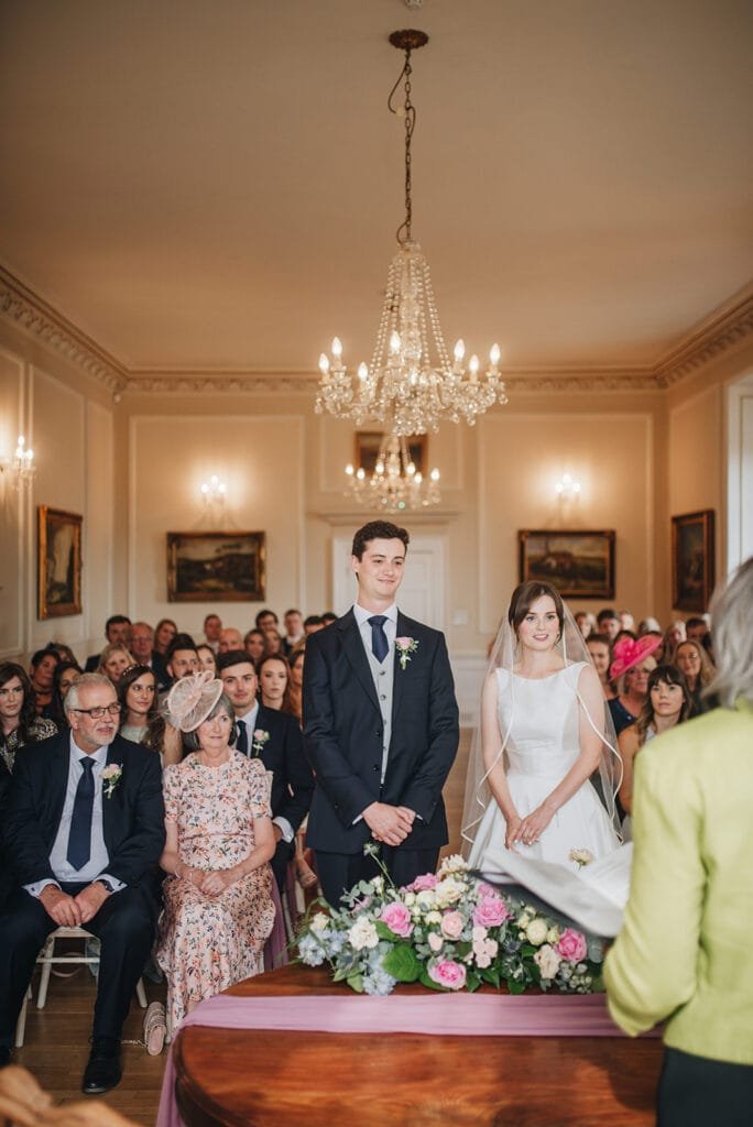 A wedding ceremony at Goldsborough Hall features a bride in a white gown and veil beside the groom in a dark suit. Guests, captured by the wedding photographers lens, admire the elegant room adorned with chandeliers, framed art, and a floral arrangement on the table.