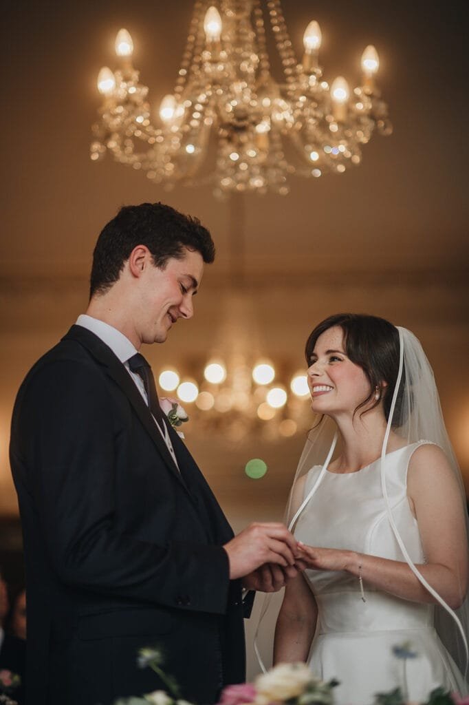 A bride and groom exchange rings beneath an elegant, lit chandelier during their wedding ceremony at Goldsborough Hall. They smile at each other as the bride wears a white dress and veil, and the groom is in a dark suit, capturing a perfect moment for any wedding photographer.