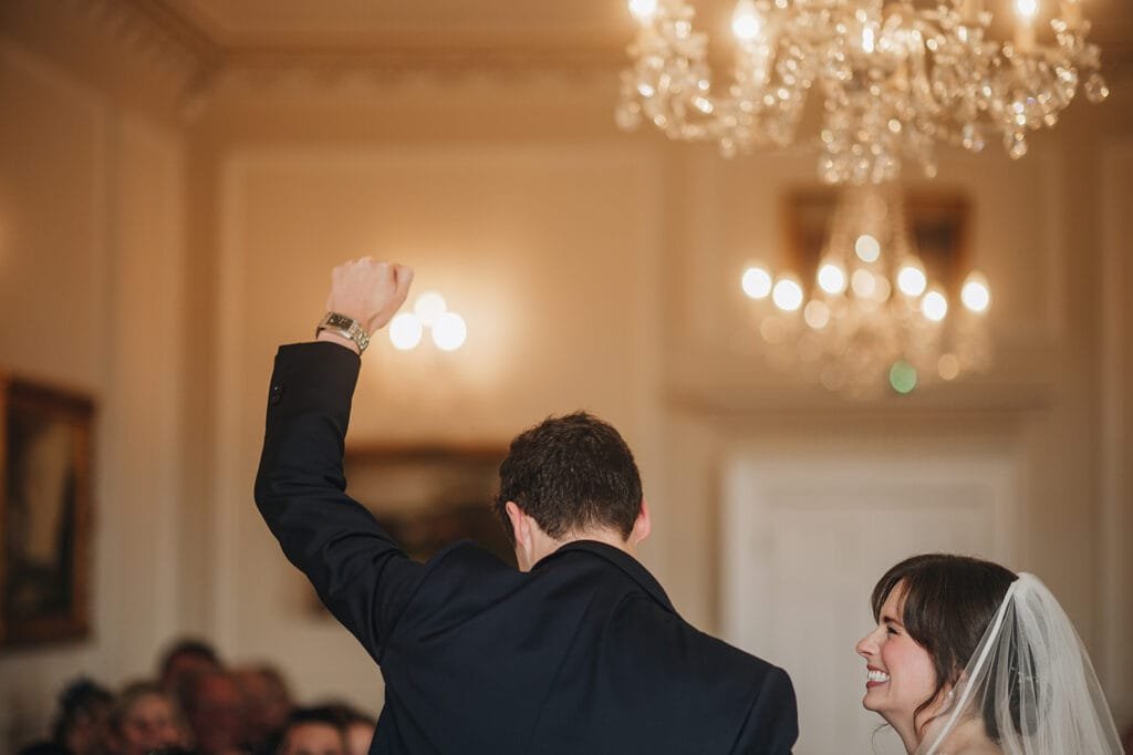 A bride and groom celebrate indoors, with the groom raising his fist in joy. The bride smiles while looking up at him. Under the glow of a chandelier at Goldsborough Hall, a wedding photographer captures this moment as a crowd sits in the softly lit room.