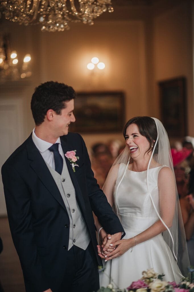 A bride and groom smile joyfully at each other during a Goldsborough Hall wedding ceremony. The bride is in a white dress and veil, while the groom sports a dark suit with a pink boutonniere. Holding hands beneath the chandeliers, their moment is perfectly captured by the wedding photographer.