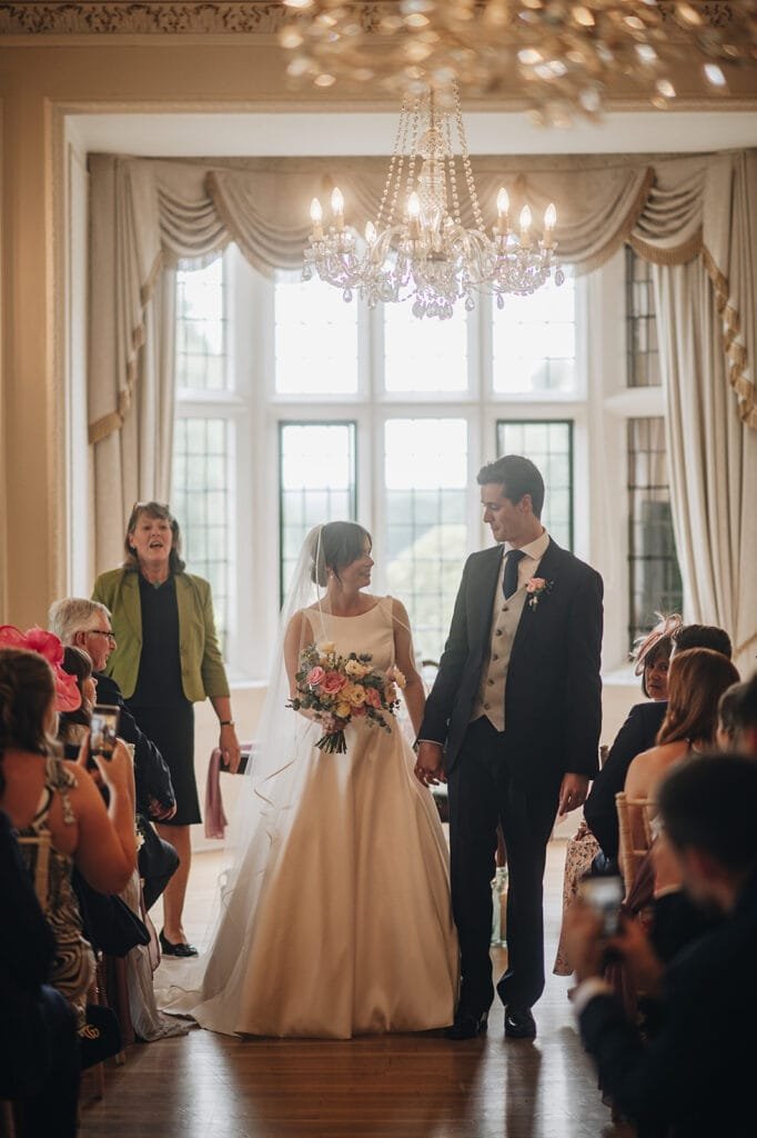 A bride and groom, holding hands, stand facing each other in a well-lit room at Goldsborough Hall with elegant drapes and a chandelier. The bride, in her white gown holding a bouquet, and the groom in his suit exchange vows as guests capture the moment on their phones.