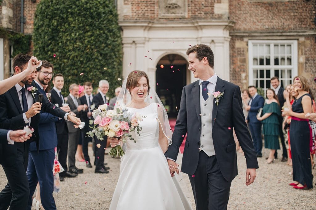 A joyful bride and groom exit a historic brick building, hand in hand, while guests cheer and throw confetti. The bride holds a bouquet of pink and white flowers, both smiling brightly. Capturing this moment is your skilled Goldsborough Hall wedding photographer amid guests in formal attire.