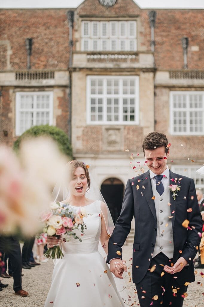 A joyful couple in wedding attire walk hand in hand outside a brick building. The bride, holding a bouquet, wears a white dress and veil, while the groom is in a dark suit. Captured by an expert Goldsborough Hall wedding photographer, they smile as petals are showered over them.