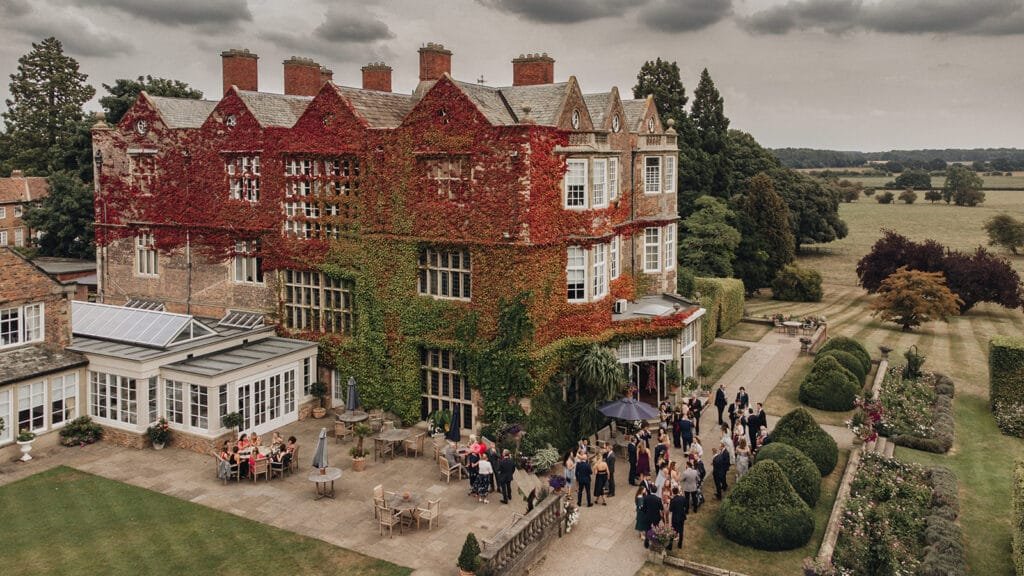 A large, ivy-covered brick building hosts a gathering on its patio. People are mingling beside tables with umbrellas, surrounded by manicured gardens, lawns, and hedges. An overcast sky adds a dramatic backdrop—perfect for a Goldsborough Hall wedding photographer to capture unforgettable moments.