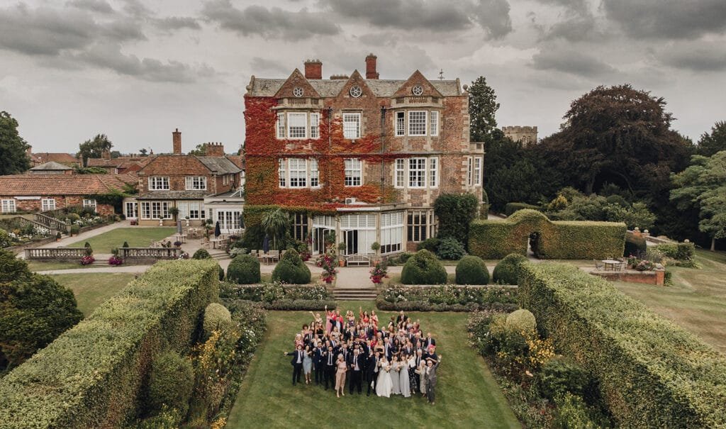 A group of people gathered for a wedding celebration in front of an ivy-covered historic mansion. Captured by a Goldsborough Hall wedding photographer, the scene is surrounded by manicured gardens and hedges, under a cloudy sky.