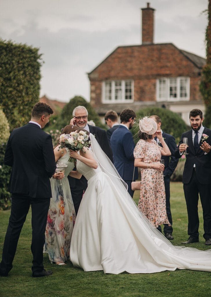 At a Goldsborough Hall wedding, a bride in a white gown and veil leans forward to hug a man holding a bouquet. Guests in formal attire, some clutching drinks, gather nearby against the backdrop of the elegant house and garden, with cloudy skies casting a soft light over the moment.