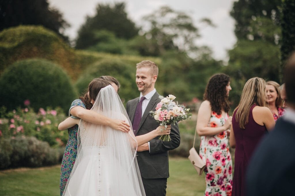 In a picturesque garden setting, the bride in her white gown and veil embraces a guest in a floral dress. Nearby, a man in a suit, holding a bouquet, smiles warmly. Other guests, draped in colorful attire, mingle amidst the greenery. A Goldsborough Hall wedding photographer captures each precious moment.