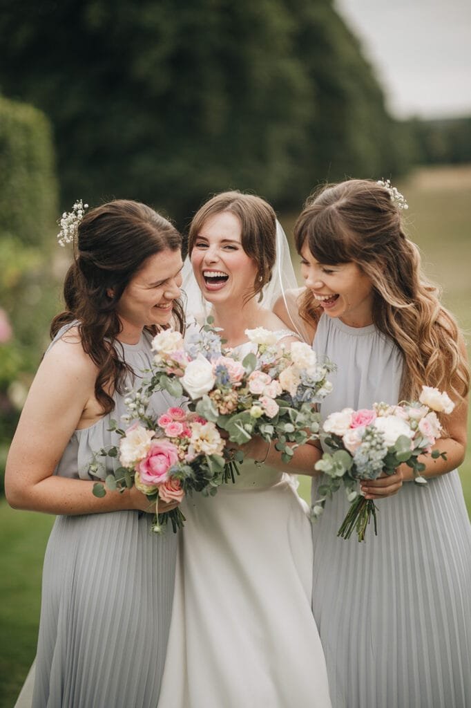 A bride in a white dress and veil laughs joyfully with two bridesmaids in gray at Goldsborough Hall. Each holds a bouquet of pink, white, and blue flowers as they stand outdoors, surrounded by lush greenery, perfectly captured by the wedding photographer.