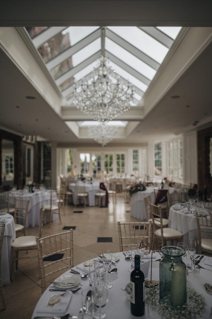 Elegant wedding reception venue with round tables draped in white cloth, set with glasses and cutlery. A large chandelier hangs from a glass-paneled ceiling, and natural light fills the room. With chairs arranged neatly around each table, its a perfect setting for capturing timeless moments by a Goldsborough Hall wedding photographer.