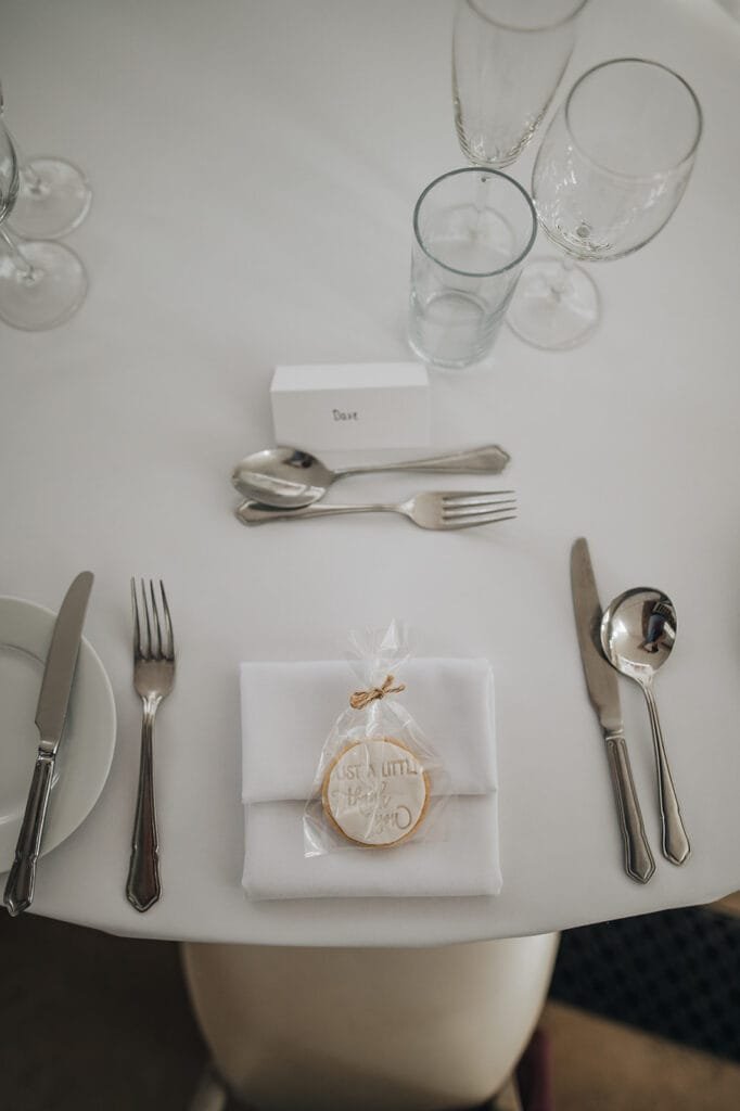 A table setting with a white plate, silverware, and a wine glass exudes elegance. At its heart, a napkin cradles a small gift wrapped in clear plastic labeled Just A Little Happy. A name card reads Dave, capturing the charm only a Goldsborough Hall wedding photographer could immortalize.