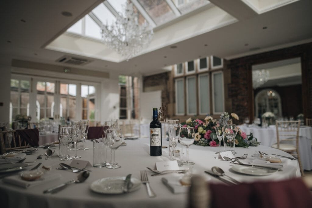 A beautifully set dining table in a bright, elegant room with a skylight evokes the charm captured by a Goldsborough Hall wedding photographer. The table features glassware, wine, and a floral centerpiece, with chairs arranged around it as soft natural light fills the space.