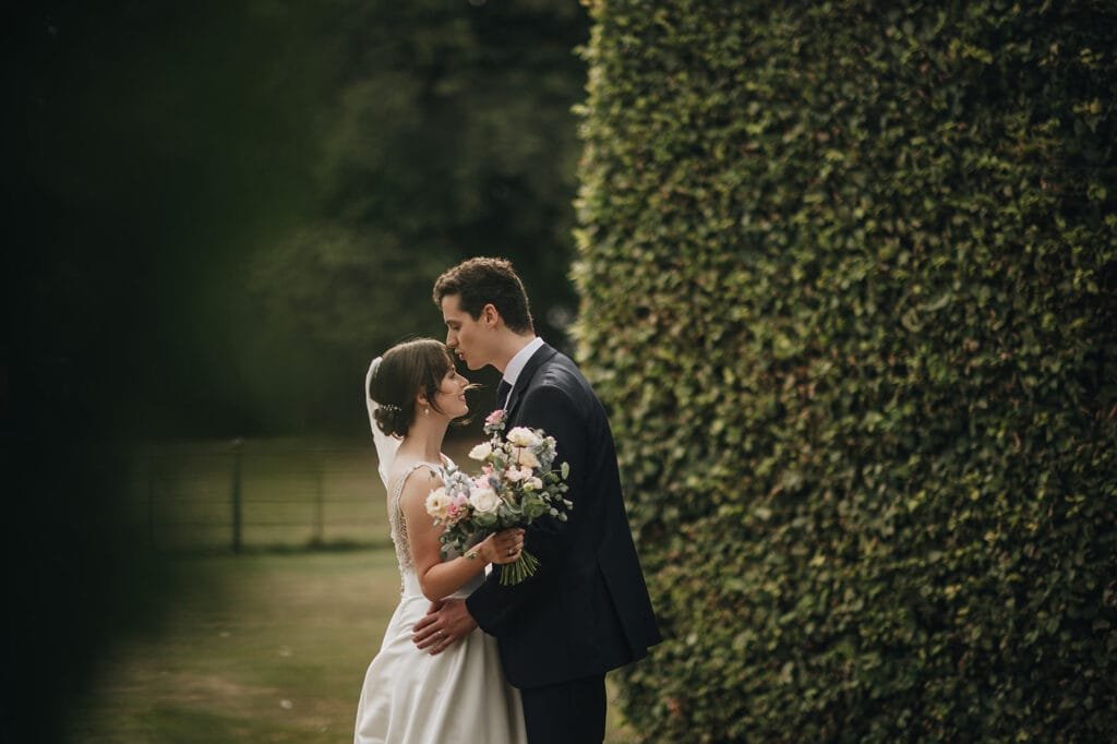 A bride and groom stand closely beside a tall hedge at Goldsborough Hall. The groom kisses the brides forehead as she holds a bouquet of flowers. They are both in wedding attire, captured beautifully by their wedding photographer against the lush garden setting.