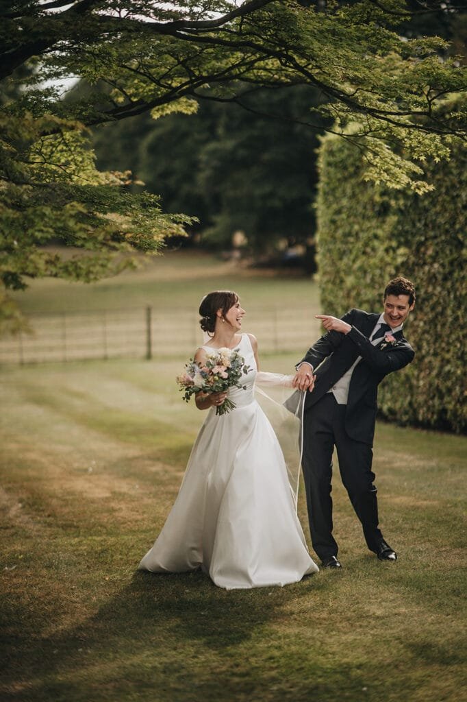 At Goldsborough Hall, a bride and groom joyously pose under a grand tree in the grassy garden, surrounded by neatly trimmed hedges. The brides laughter fills the air as she holds her bouquet while the groom playfully points away, both appearing happy and relaxed.