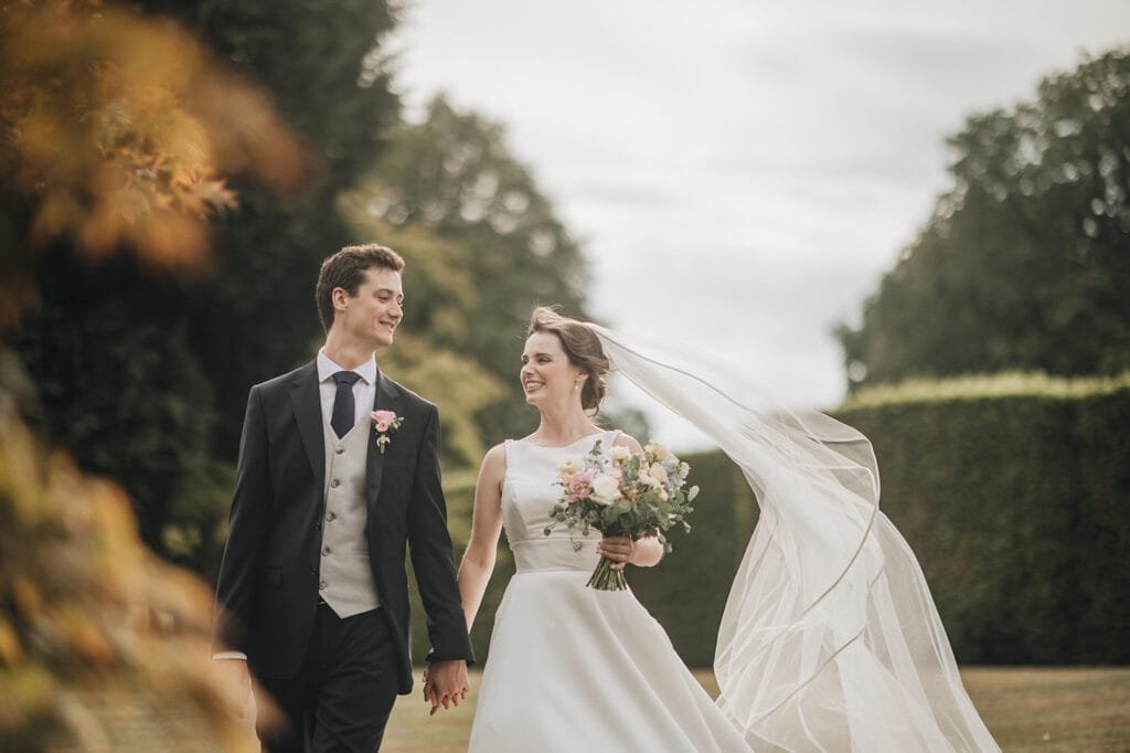 A bride and groom, smiling and holding hands, walk outside at Goldsborough Hall. The brides white dress with a long veil flows in the wind as she holds a bouquet. The groom is in a dark suit and vest. Greenery and blurred trees create a serene backdrop for this magical moment captured by their wedding photographer.