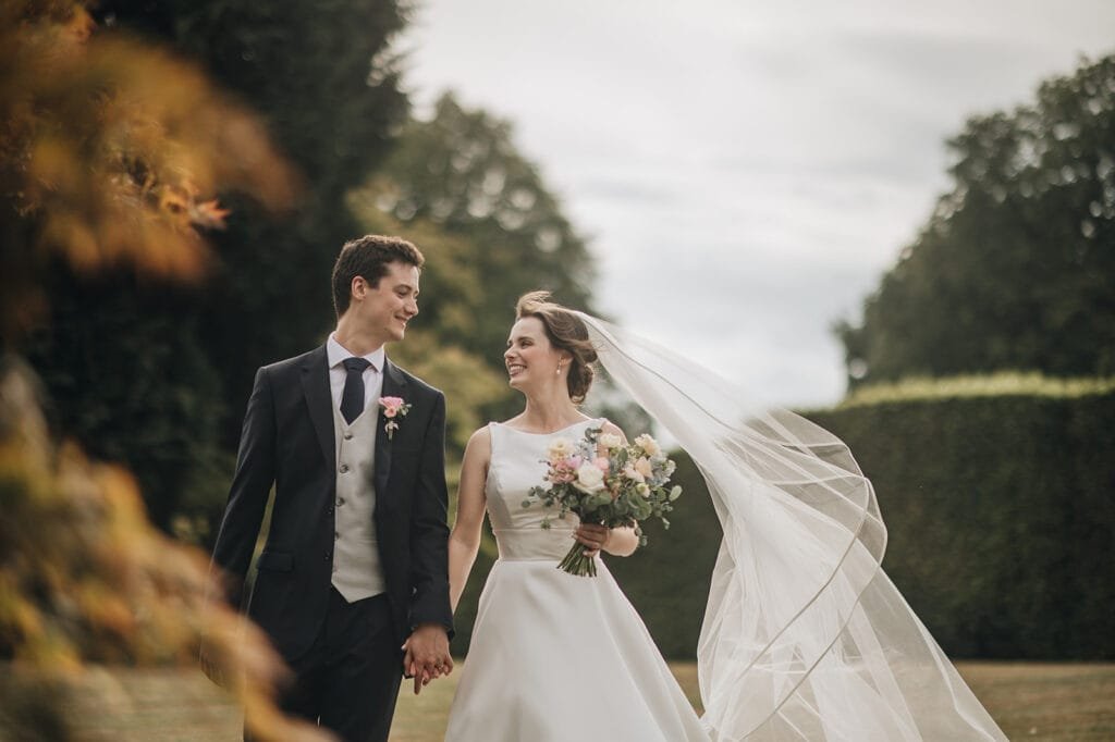 A bride and groom walk hand in hand outdoors at Goldsborough Hall. The bride, in a white dress and veil, holds a bouquet of flowers. The groom dons a dark suit with vest and tie. Smiling at each other, they are surrounded by lush greenery—a perfect wedding photo moment.