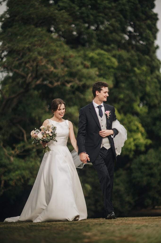 A bride and groom walk hand in hand on a grassy path at Goldsborough Hall. The bride, in a white gown holding a bouquet, and the groom, in a dark suit with a tie, appear happy amidst lush greenery. Its a picture-perfect moment captured by their wedding photographer.