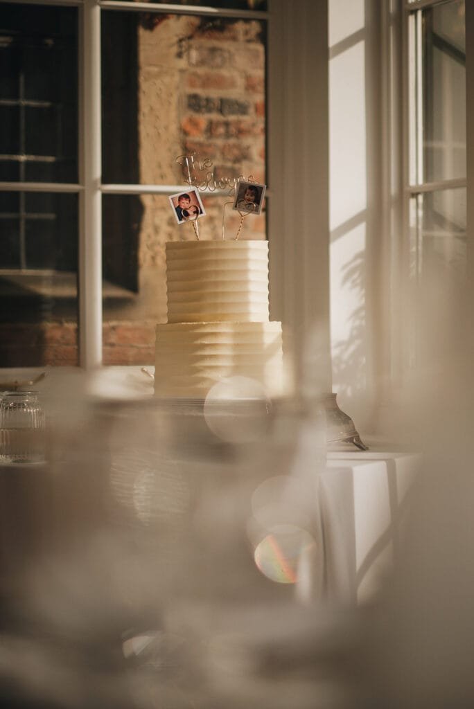 A tiered white cake with ribbed texture is elegantly showcased in a sunlit room, perfect for any Goldsborough Hall wedding photographer to capture. It features photo toppers and a script word decoration, while a window reveals a glimpse of the brick wall outside. Blurred elements frame this exquisite scene.