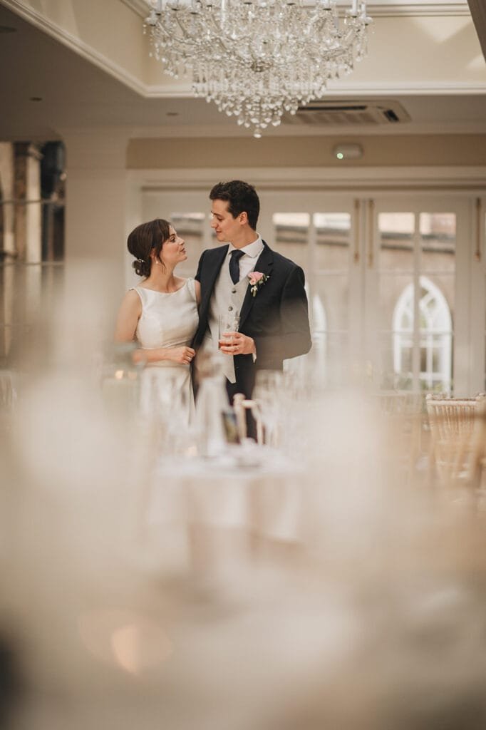 A bride and groom smile at each other in an elegant Goldsborough Hall wedding. The bride wears a white dress, and the groom is in a suit with a tie. A chandelier hangs above while a skilled wedding photographer captures every moment amidst beautifully set tables in the background.