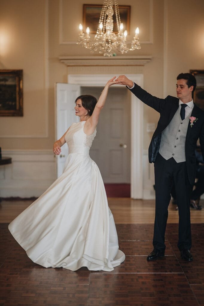 A bride in a flowing white wedding dress and a groom in a dark suit dance gracefully at Goldsborough Hall, their elegance mirrored by chandeliers and framed paintings. As she twirls, the moment is perfectly captured by a skilled wedding photographer.