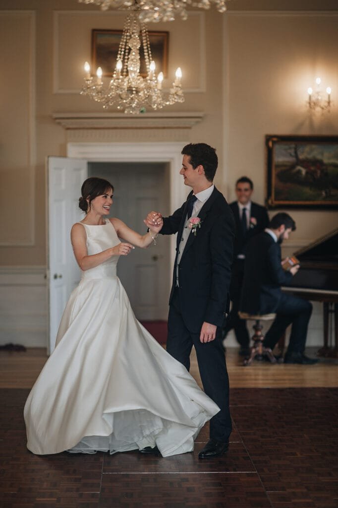 A bride in a white gown and a groom in a black suit joyfully dance beneath the chandelier at Goldsborough Hall. A man in a suit plays the piano softly, capturing the elegance and warmth of this enchanting moment through their wedding photographers lens.