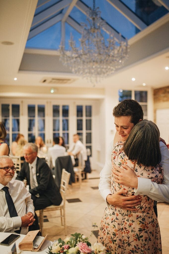 A young man and an older woman embrace warmly in a well-lit, elegant room with a glass ceiling and chandelier, beautifully captured by the Goldsborough Hall wedding photographer. Other guests sit and chat at tables in the background.