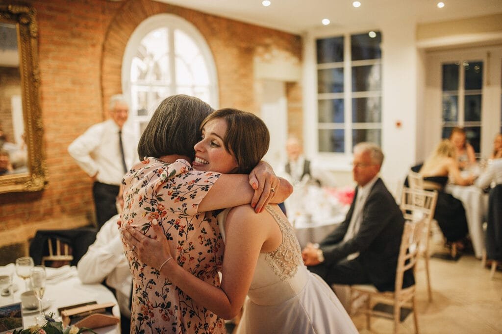 A woman in a white dress happily hugs another woman in a floral blouse at a Goldsborough Hall wedding, warmly lit with guests seated at tables nearby. A large arched window and mirror adorn the brick wall, capturing the joyous moments perfectly.