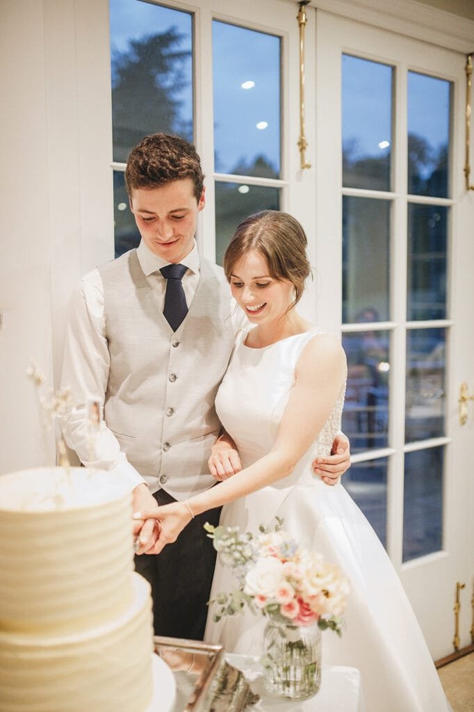 At a picturesque Goldsborough Hall wedding, the bride and groom joyfully cut their tiered cake. She dazzles in white, while he sports a vest and tie. A vibrant bouquet graces the table as sunlight streams through the glass doors behind them, all beautifully captured by their wedding photographer.